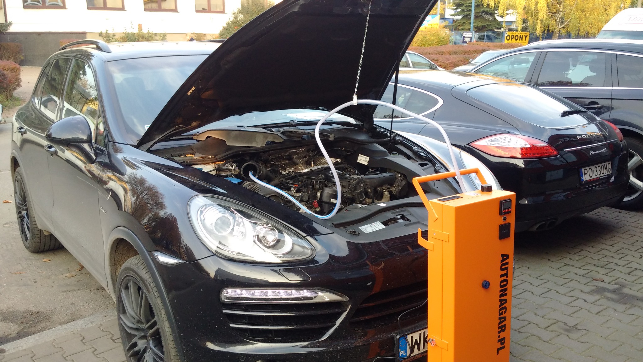 Czarny Porsche Cayenne z otwartą maską, podłączony do urządzenia do wodorowania silnika Autonagar.pl, na tle widoczny drugi Porsche Panamera.