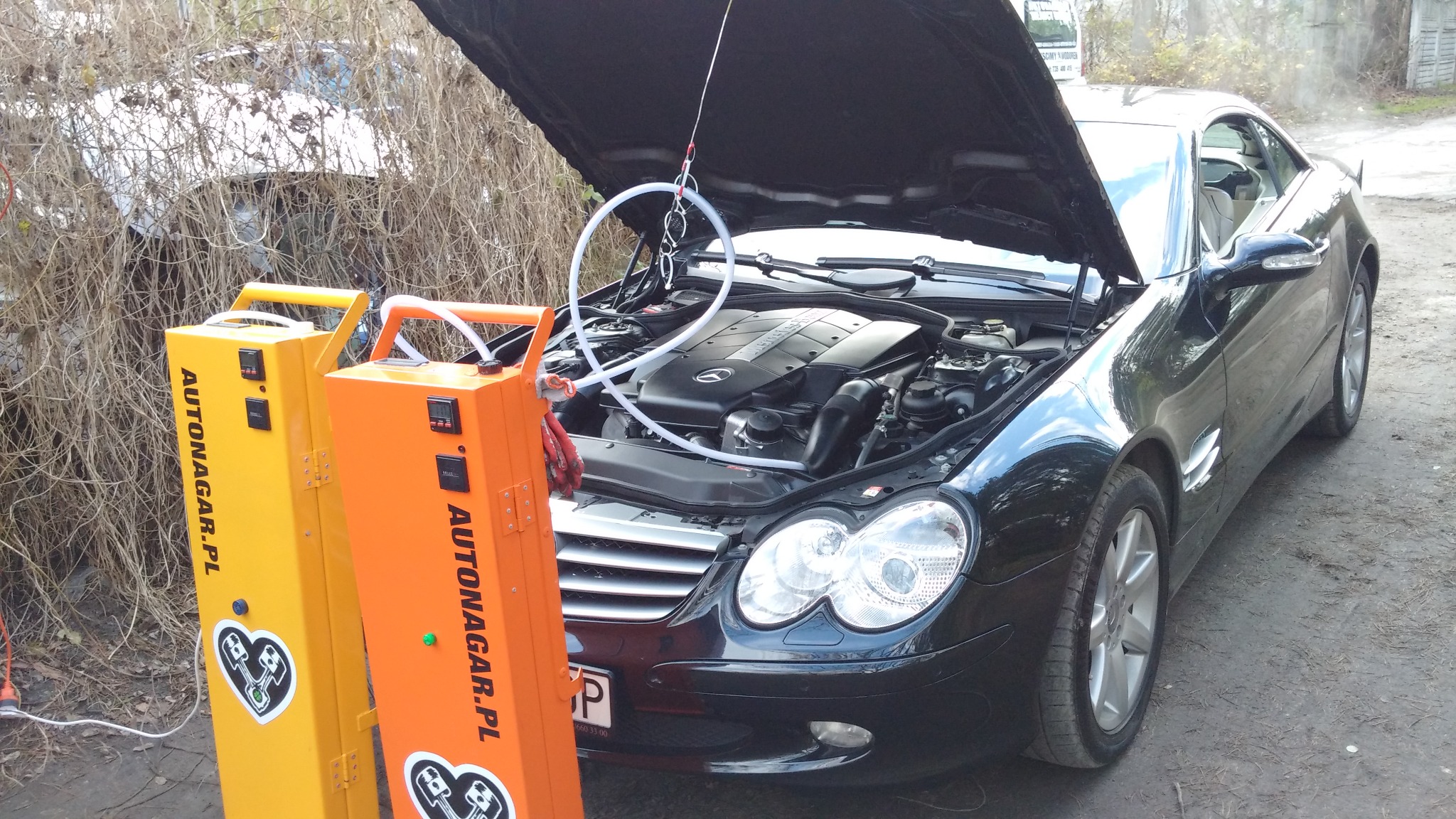 Czarny Mercedes SLC z otwartą maską silnika, podłączony do urządzenia do wodorowania silnika Autonagar.pl, widoczne żółte i pomarańczowe obudowy urządzenia, tło zarośnięte krzakami.