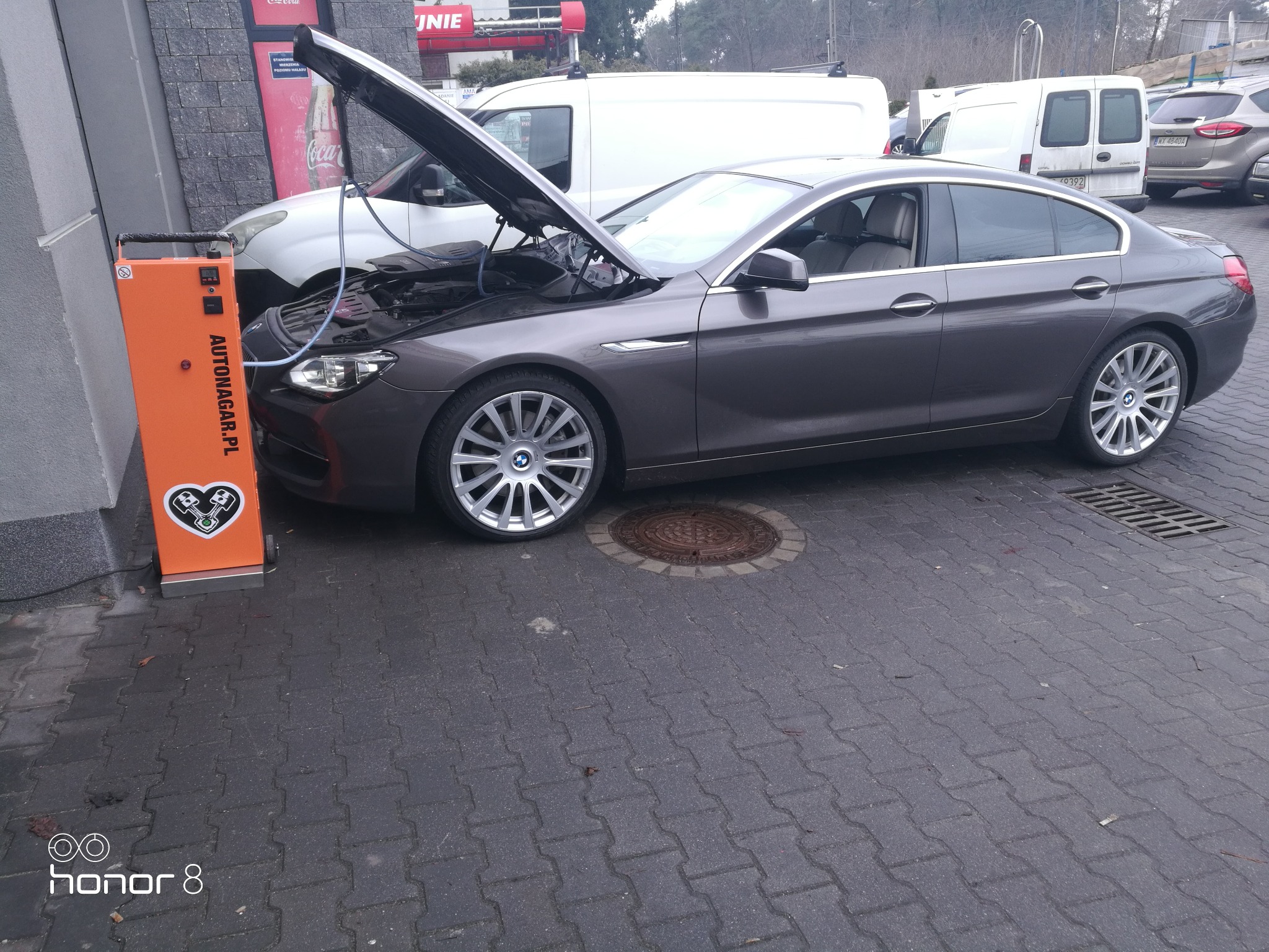 Szary samochód BMW z otwartą maską podłączony do pomarańczowego urządzenia do wodorowania silnika Autonagar.pl na zewnątrz warsztatu.