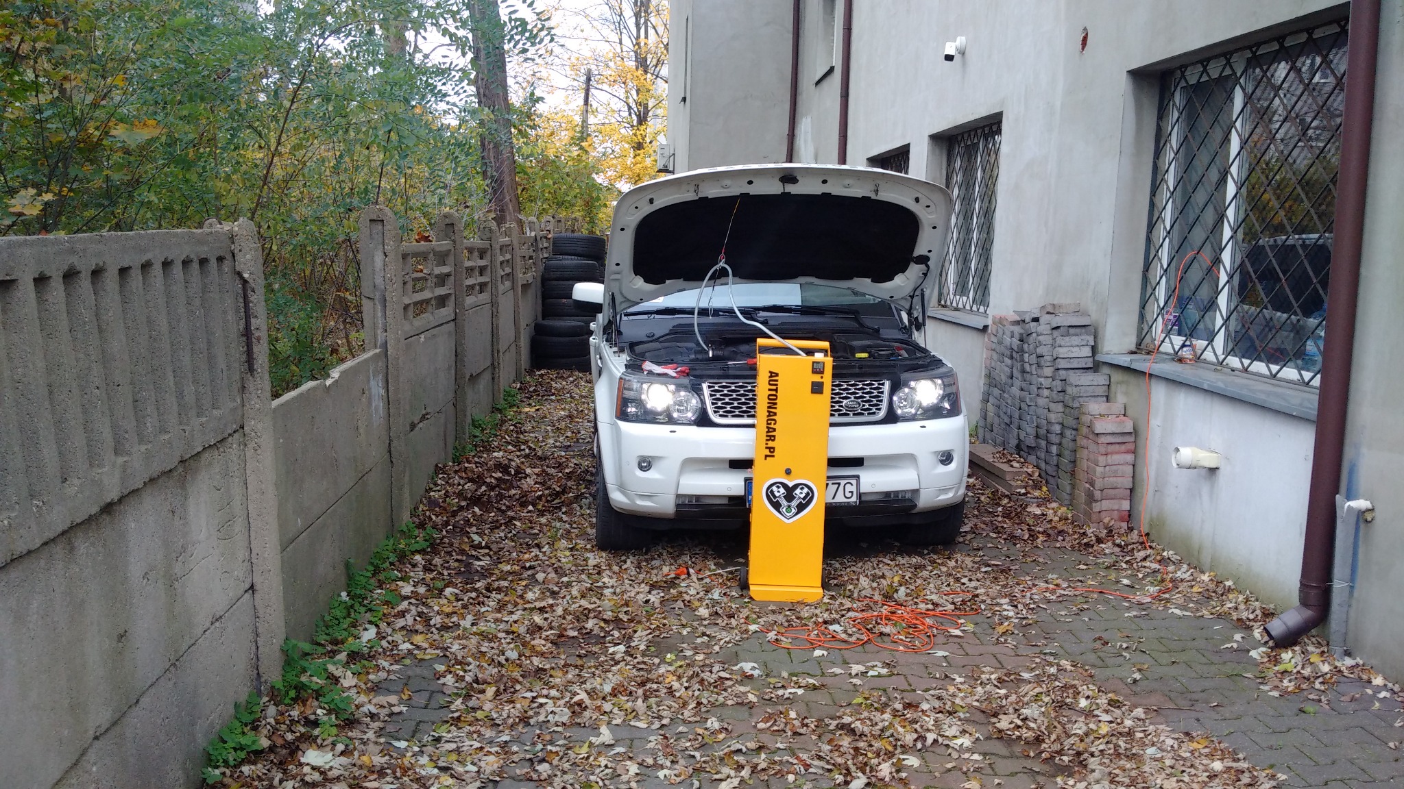 Biały Range Rover z otwartą maską, podłączony do żółtego urządzenia do wodorowania silnika Autonagar.pl, na podwórku z liśćmi.