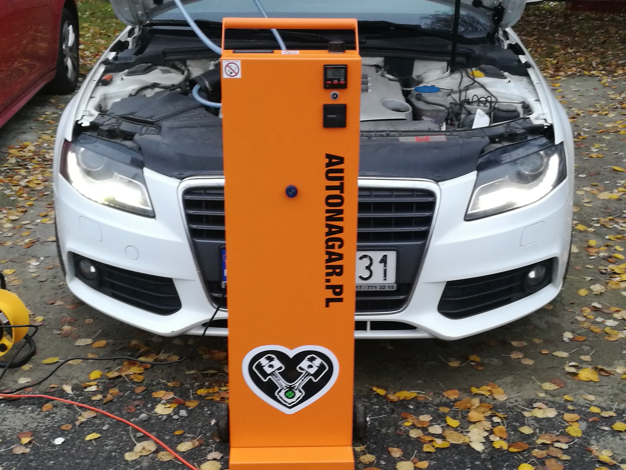 Białe auto z otwartą maską podczas wodorowania silnika za pomocą pomarańczowego urządzenia Autonagar.pl z logiem serca i tłoków na tle jesiennych liści.