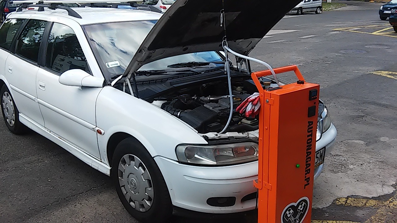 Biały Opel kombi z otwartą maską podczas procesu wodorowania silnika, widoczna pomarańczowa maszyna do wodorowania Autonagar.pl z podłączonymi przewodami.
