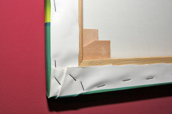 Fragment naciągniętego płótna na drewnianej ramie, widoczne zszywki mocujące i narożnik z klinami, na tle różowej ściany.