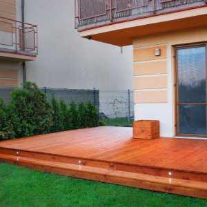 Drewniany taras z oświetleniem punktowym wbudowanym w stopień, przylegający do jasnego budynku z balkonem i dekoracyjnymi pasami elewacyjnymi, otoczony trawnikiem i tujami.