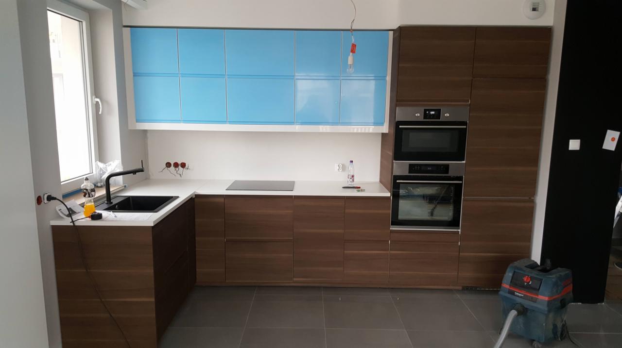 Nowoczesna kuchnia z brązowymi szafkami dolnymi i błękitnymi górnymi, biały blat, czarny zlew i płyta indukcyjna, widoczny odkurzacz przemysłowy na szarej podłodze.