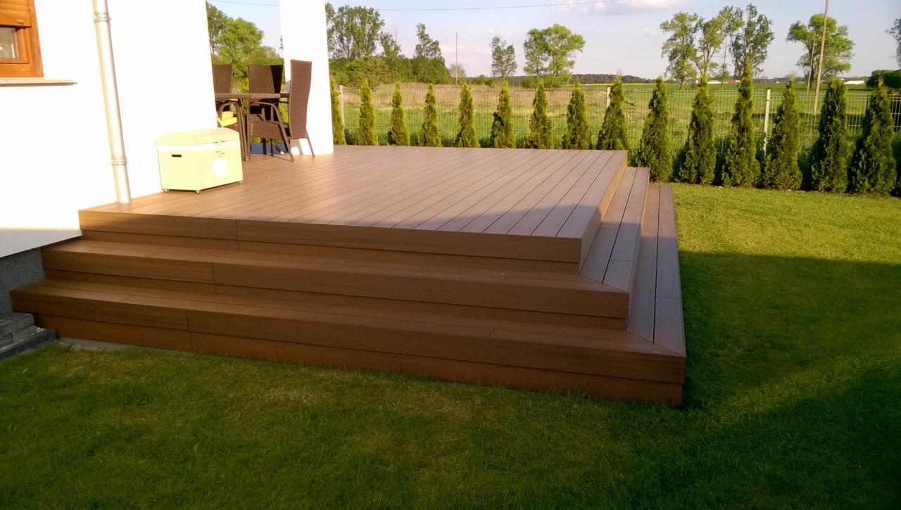 Drewniany taras z trzema schodkami, przylegający do białego budynku, z widocznymi meblami ogrodowymi na tarasie i żywopłotem w tle.