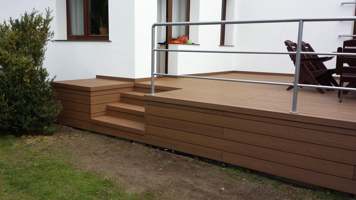 Nowoczesny taras z kompozytu w kolorze brązu, z metalową barierką, przylegający do białego budynku z brązowymi oknami, widok z boku na schody i krzesła ogrodowe.