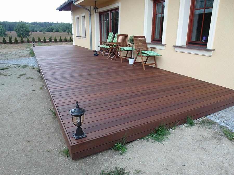 Rozległy taras z ciemnego drewna egzotycznego przylegający do domu z beżową elewacją, z widocznymi drewnianymi krzesłami ogrodowymi i lampą stojącą przy krawędzi tarasu.