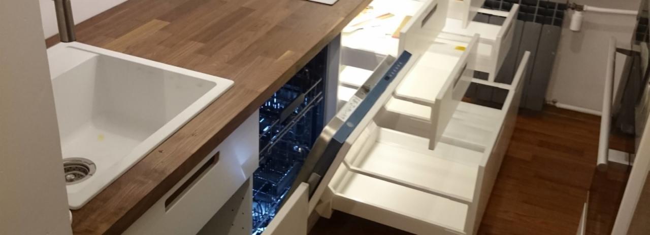 Nowoczesna kuchnia z drewnianym blatem, białym zlewem i otwartą zmywarką z niebieskim podświetleniem, obok białe szuflady bez uchwytów.