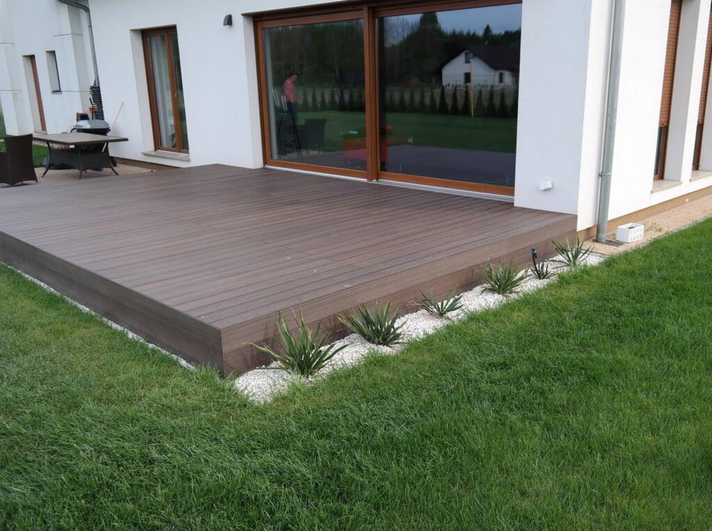 Nowoczesny taras z kompozytowych desek w kolorze ciemnego brązu, obrzeżony białym żwirem i zielonymi roślinami, przylegający do białego budynku z dużymi przesuwnymi oknami.