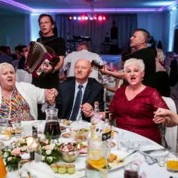 Zespół grający na weselu, akordeonista i gitarzysta umilają czas gościom siedzącym przy stole, trzymającym się za ręce i śpiewającym.