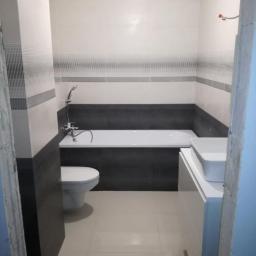 GW Construction Wykonczenia Wroclaw - Wykończona łazienka z białą armaturą, ciemnoszarą zabudową wanny i jasnymi płytkami z dekoracyjnym pasem, widok zza framugi drzwi.