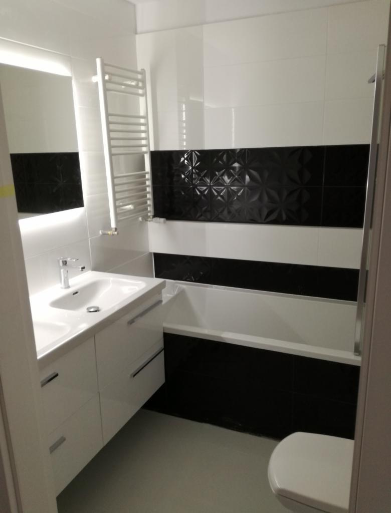 Nowoczesna łazienka z białymi szafkami pod umywalką, grzejnikiem drabinkowym i wanną obłożoną czarno-białymi płytkami z trójwymiarowym wzorem w czarnym pasie.