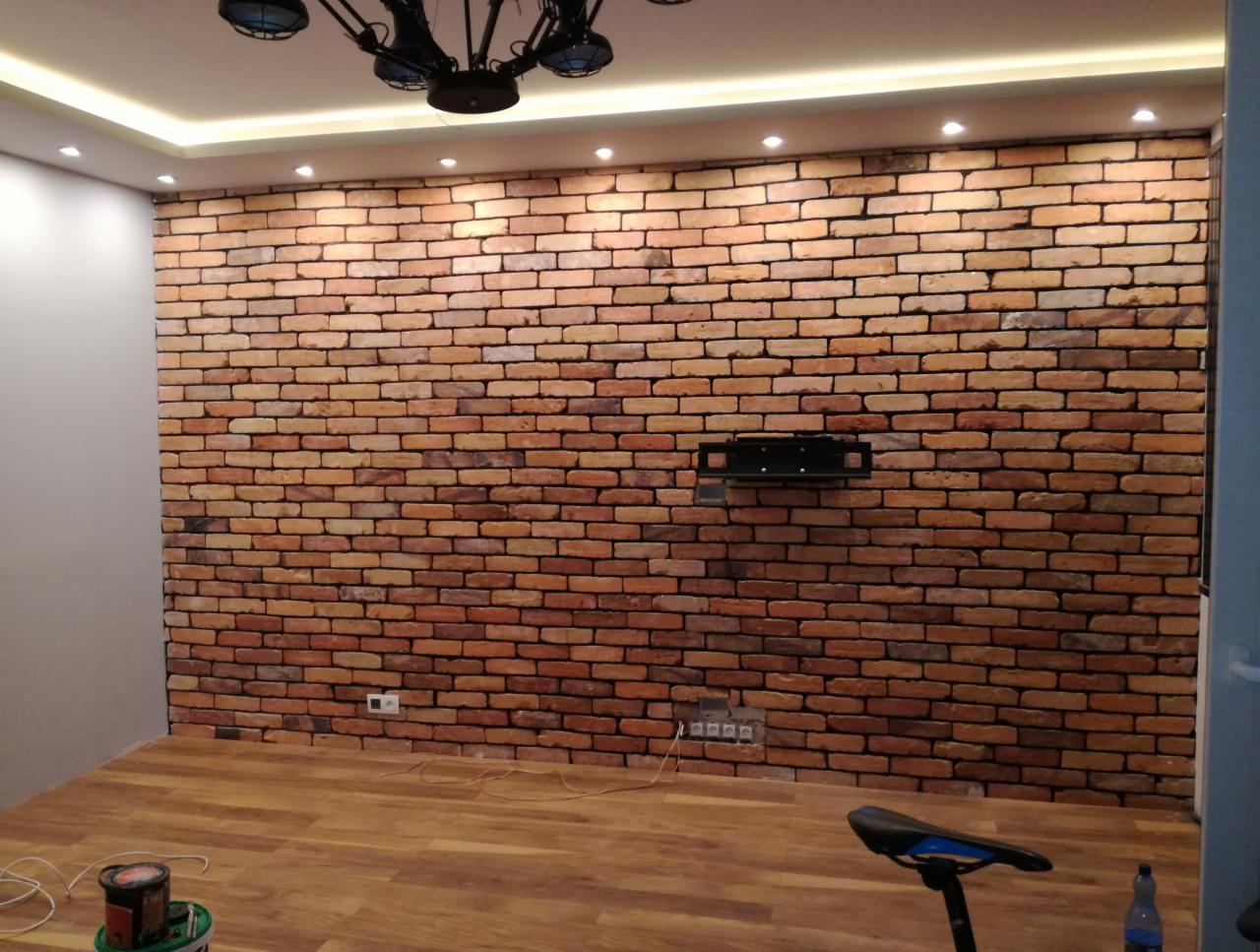 Ściana z cegły klinkierowej w salonie z zamontowanym uchwytem na telewizor, oświetlenie punktowe w suficie podwieszanym, podłoga z paneli imitujących drewno, widoczny fragment roweru stacjonarnego...
