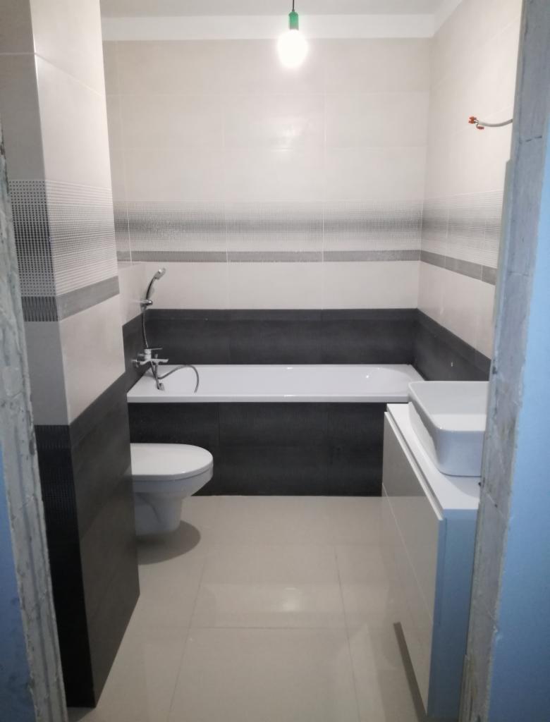 Wykończona łazienka z białą armaturą, ciemnoszarą zabudową wanny i jasnymi płytkami z dekoracyjnym pasem, widok zza framugi drzwi.