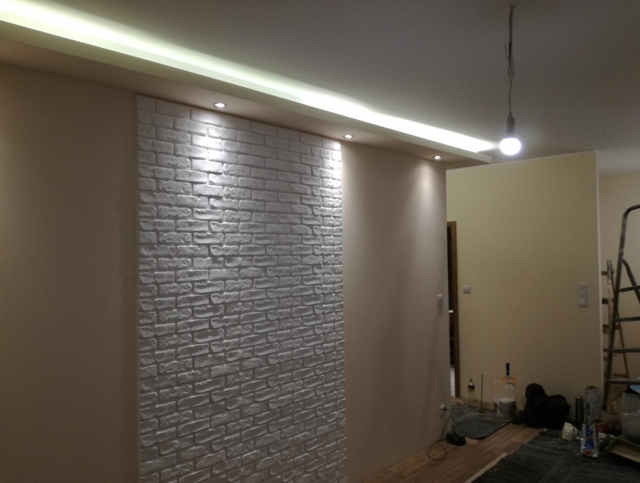 Ściana z dekoracyjnej białej cegły w salonie, z oświetleniem LED podsufitowym i pojedynczą żarówką zwisającą z sufitu. Widoczne ślady po pracach wykończeniowych na podłodze.