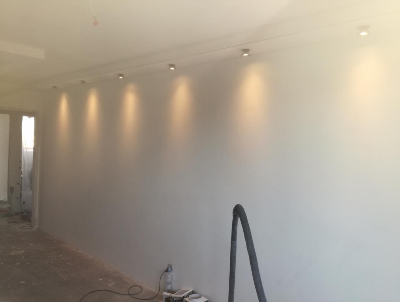 Świeżo pomalowane wnętrze z rzędem punktowych lamp sufitowych, widoczny wąż odkurzacza i butelka wody na podłodze.