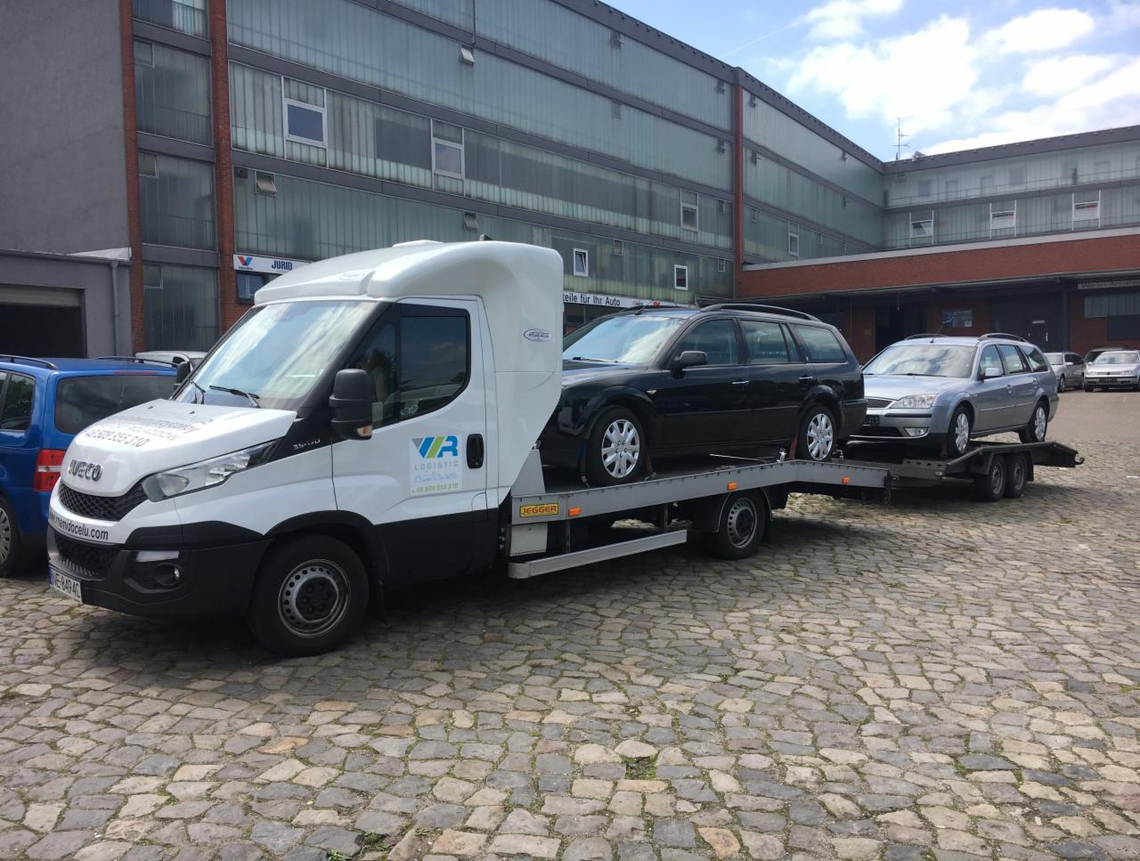 Biała laweta Iveco z logo firmy VR Logistic przewozi dwa samochody kombi: czarny i srebrny, na tle budynku z cegły i szkła.