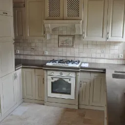 Kuchnia w stylu klasycznym z kremowymi szafkami z frezowanymi frontami, ciemnoszary blat, płytki ceramiczne na ścianie z dekoracyjnym motywem nad kuchenką gazową, oraz beżowa podłoga z płytek.