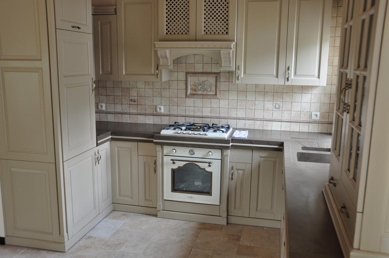 Kuchnia w stylu klasycznym z kremowymi szafkami z frezowanymi frontami, ciemnoszary blat, płytki ceramiczne na ścianie z dekoracyjnym motywem nad kuchenką gazową, oraz beżowa podłoga z płytek.