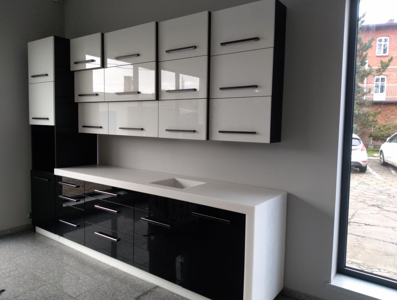 Czarno-biała zabudowa kuchenna z błyszczącymi frontami i długimi, poziomymi uchwytami, widoczny zlew wpuszczony w biały blat, na tle okna z widokiem na budynek i zaparkowane samochody.