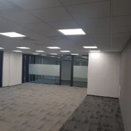 Puste, jasne biuro z szarą wykładziną dywanową w geometryczny wzór, szklane ścianki działowe z matową folią na wysokości wzroku, białe ściany i sufit z panelami LED.