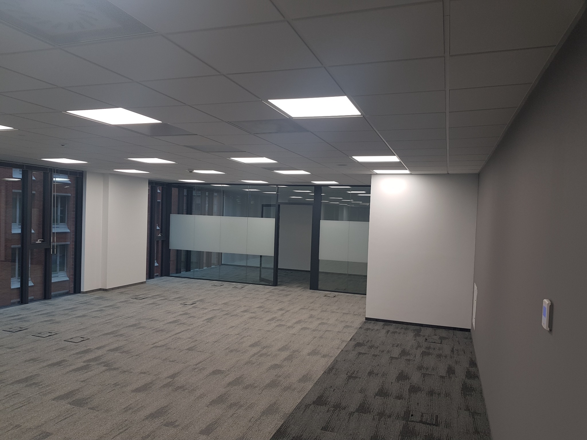 Puste, jasne biuro z szarą wykładziną dywanową w geometryczny wzór, szklane ścianki działowe z matową folią na wysokości wzroku, białe ściany i sufit z panelami LED.