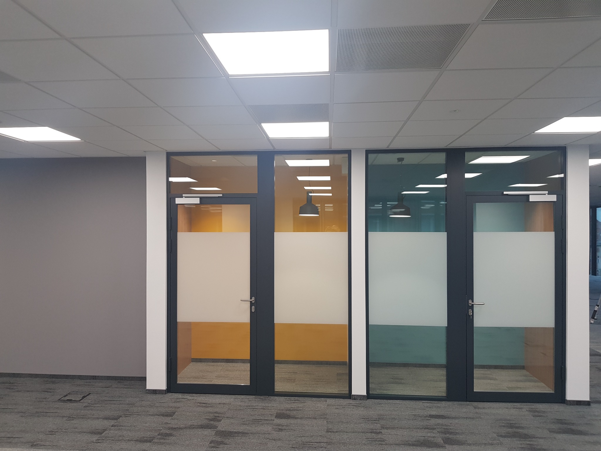 Przeszklone drzwi biurowe z matową folią w nowocześnie wykończonym wnętrzu z podwieszanym sufitem i szarą wykładziną.