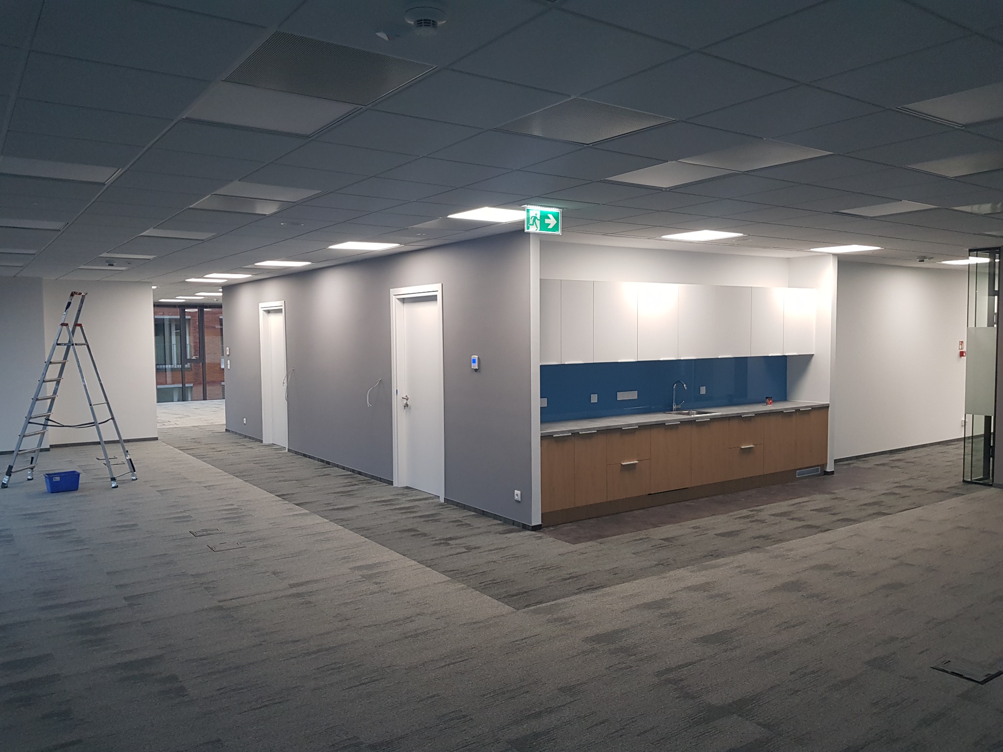 Wnętrze biura po remoncie z szarą wykładziną dywanową, świeżo pomalowanymi szarymi ścianami, białą zabudową kuchenną z niebieskim panelem i drabiną malarską w rogu.