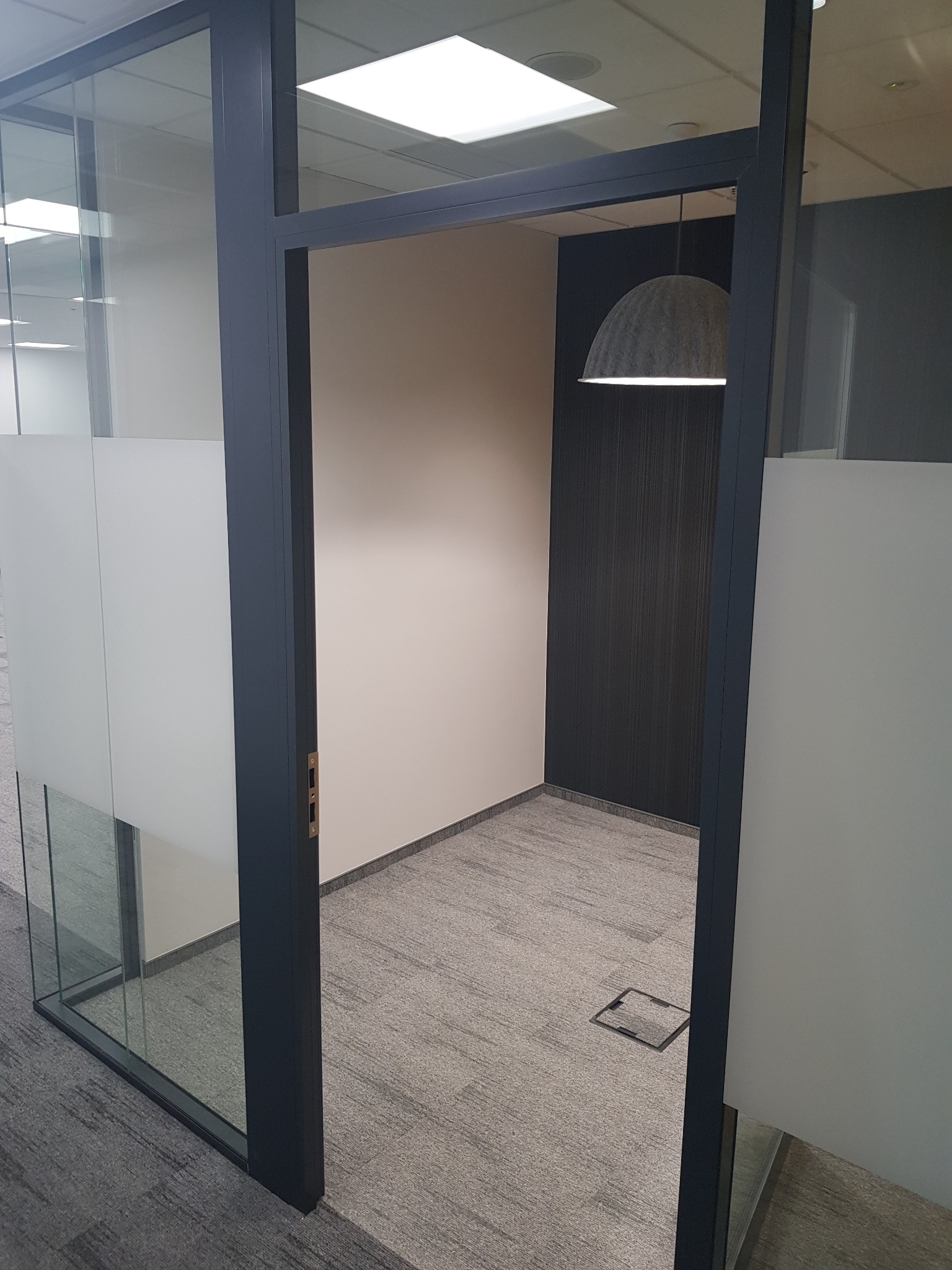 Nowoczesne biuro z przeszkloną ścianą i drzwiami, widoczny szary dywan, białe ściany i designerska lampa wisząca w kształcie kopuły.