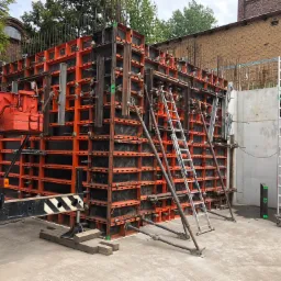 Szczegółowe ujęcie szalunku fundamentowego z widocznym żurawiem, stalowymi prętami zbrojeniowymi i aluminiową drabiną na placu budowy, z budynkiem z czerwonej cegły w tle.