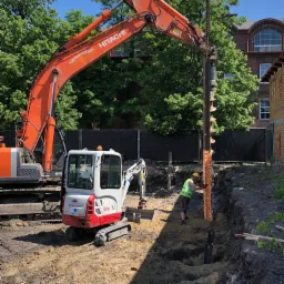 Pomarańczowa koparka Hitachi i biała mini koparka Takeuchi TB216 na placu budowy podczas wiercenia otworów pod fundamenty z pomocą wiertła ślimakowego i pracownika w kasku i kamizelce odblaskowej.