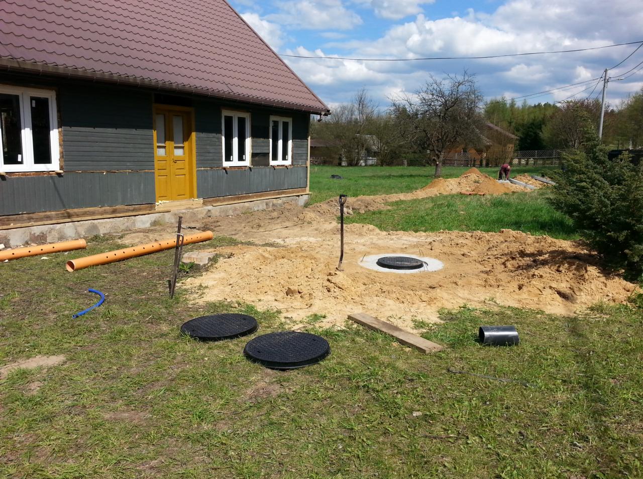 Teren po wykonanych pracach ziemnych z widocznymi elementami studni kanalizacyjnej, rurami PCV i wykopem obok drewnianego domu z żółtymi drzwiami.