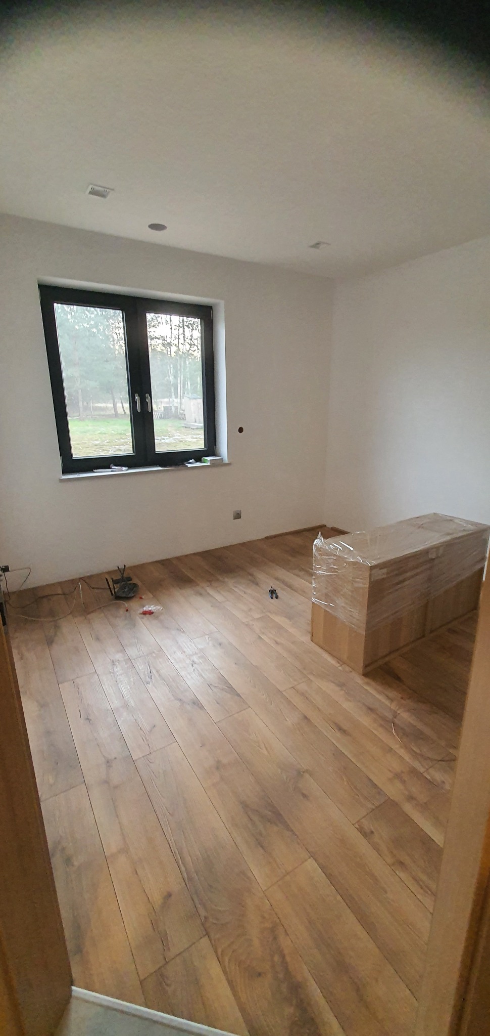 Nowo położona, błyszcząca podłoga z paneli drewnopodobnych w pustym pokoju z oknem, zapakowane panele pod ścianą, widoczne kable i małe elementy na podłodze.
