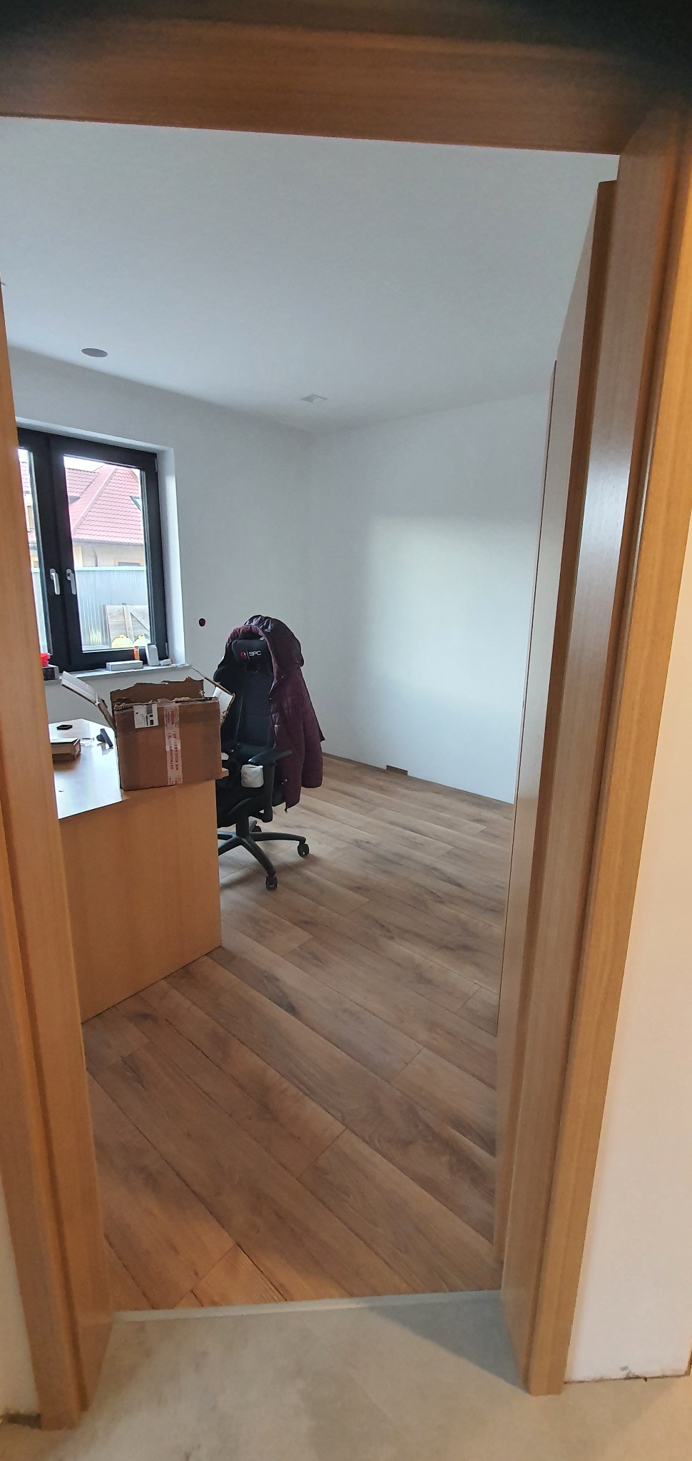Widok przez futrynę na pokój z nową drewnianą podłogą ułożoną w jodełkę, biurko z kartonem i krzesło z kurtką, ściany pomalowane na biało, widoczne ślady po pracach wykończeniowych przy podłodze.