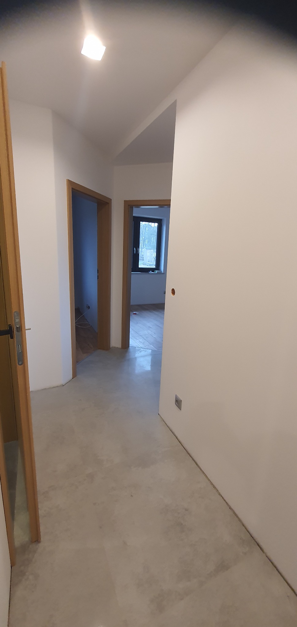 Korytarz w trakcie wykańczania, widoczne surowe ściany, wylewka na podłodze, ościeżnice drzwiowe w kolorze drewna, widok na okno w oddali.