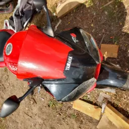 Czerwono-czarny motocykl Yamaha R1 w ujęciu z góry, zaparkowany na ziemi obok drewnianych elementów. Widoczne lusterka, kierownica i fragment opony.