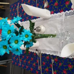 Udekorowany stół z bukietem błękitnych kwiatów w szklanym wazonie, białą świecą w szklanym świeczniku, solniczką, białym bieżnikiem z koronką, złożonymi serwetkami i sztućcami na niebieskim obrusie...