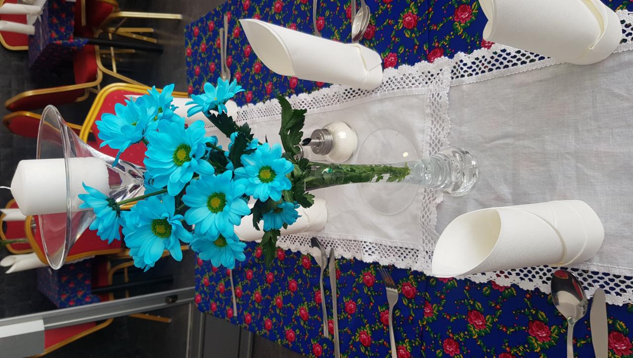 Udekorowany stół z bukietem błękitnych kwiatów w szklanym wazonie, białą świecą w szklanym świeczniku, solniczką, białym bieżnikiem z koronką, złożonymi serwetkami i sztućcami na niebieskim obrusie...