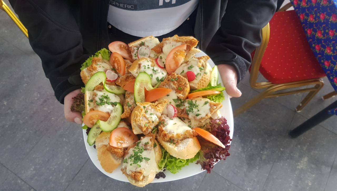 Osoba trzyma duży talerz z kanapeczkami z kotletem, serem, pomidorami, ogórkami i rzodkiewką, udekorowanymi zieleniną, na tle krzesła z czerwonym obiciem w kropki.