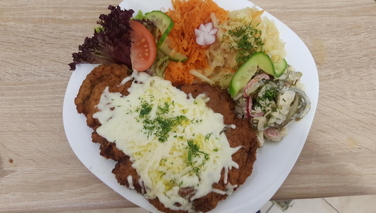 Szaszłyk wieprzowy panierowany podany z tartą marchewką, surówką z kapusty, sałatką warzywną i dekoracją z pomidora i sałaty na białym talerzu.