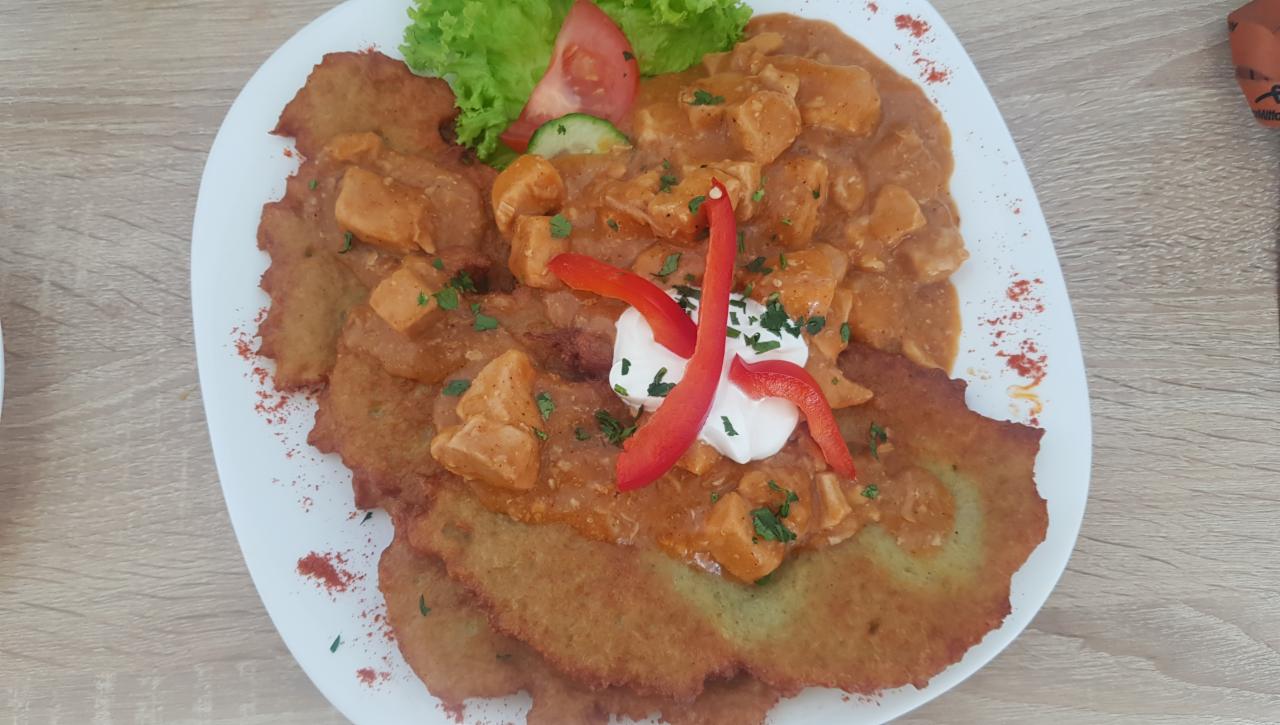 Placek ziemniaczany z gulaszem i kleksem śmietany, udekorowany papryką i zieloną pietruszką, podany na białym talerzu z dodatkiem sałaty i pomidora.