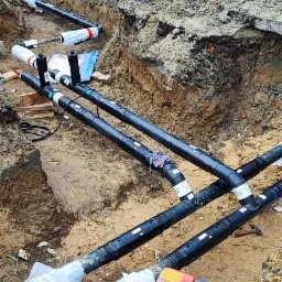 Wykop z ułożonymi czarnymi rurami z białymi etykietami i owinięciami ochronnymi, czerwona skrzynka narzędziowa w rogu, w tle budynek z ceglanymi elementami