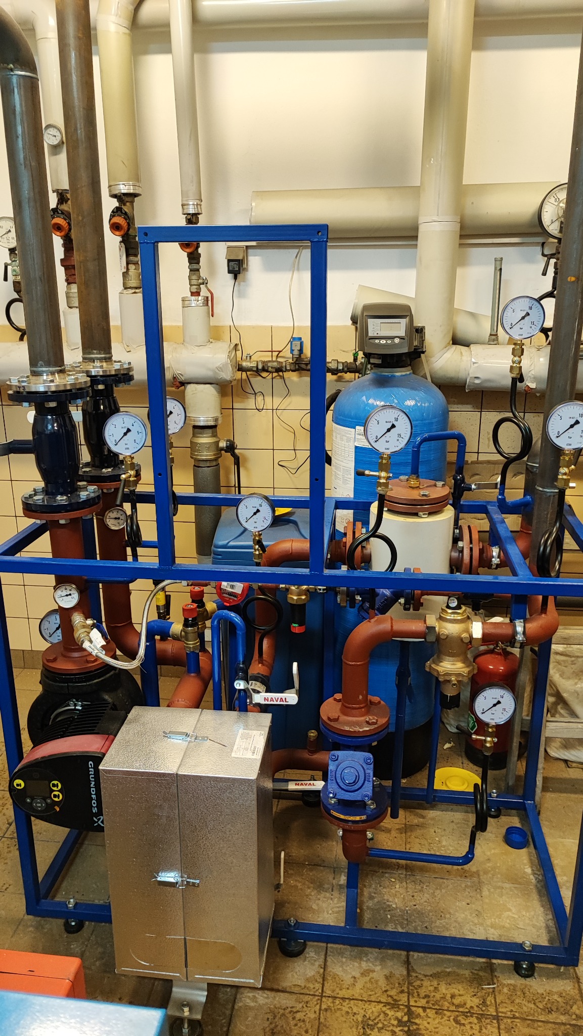 Złożony system rur, zaworów i manometrów w niebieskiej ramie, z widocznym srebrnym modułem zamykanym na zatrzaski i pompą Grundfos, na tle ściany z kafelkami.