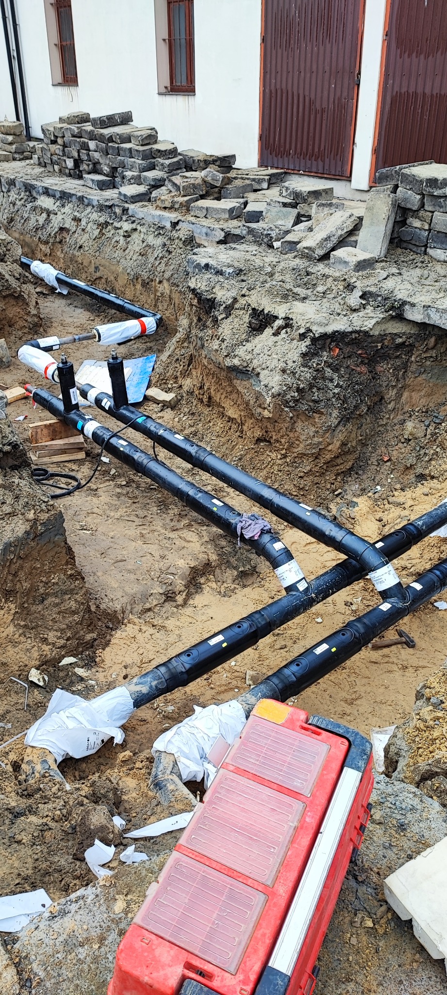 Wykop z ułożonymi czarnymi rurami z białymi etykietami i owinięciami ochronnymi, czerwona skrzynka narzędziowa w rogu, w tle budynek z ceglanymi elementami
