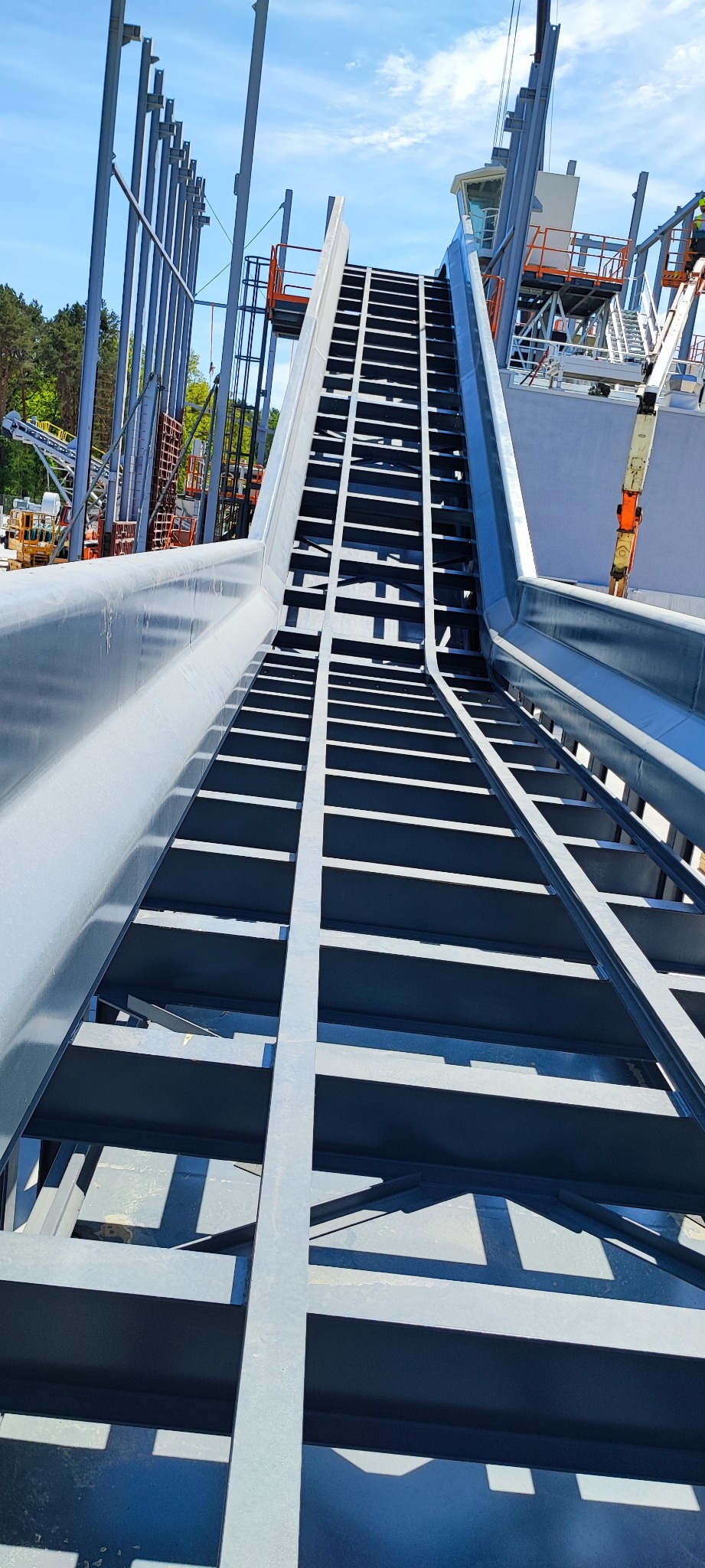 Konstrukcja stalowa pod kątem, prawdopodobnie fragment większej budowli przemysłowej, z widocznymi spawami i elementami łączeniowymi, na tle błękitnego nieba i zieleni drzew.