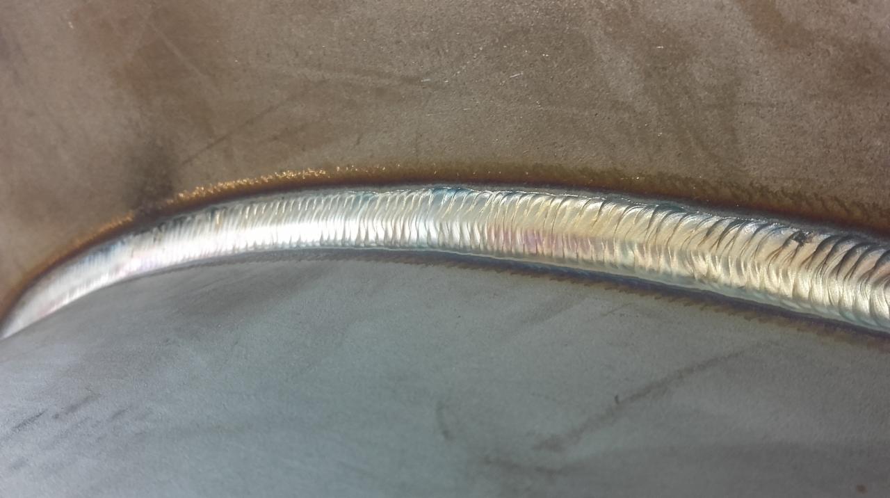 Detal spoiny na metalowej powierzchni, widoczne delikatne opalizujące kolory i struktura spoiny.