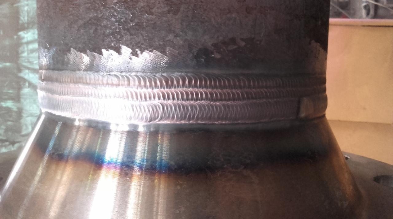 Połączenie spawane rury ze stożkowym elementem, widoczna struktura spoiny z charakterystycznym wzorem, przebarwienia termiczne metalu w strefie spawania.