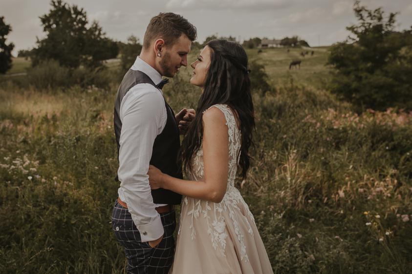 Para młoda w objęciach na łące; panna młoda w sukni z koronkowymi aplikacjami, pan młody w kamizelce i spodniach w kratę, w tle zielona łąka i pasące się konie.