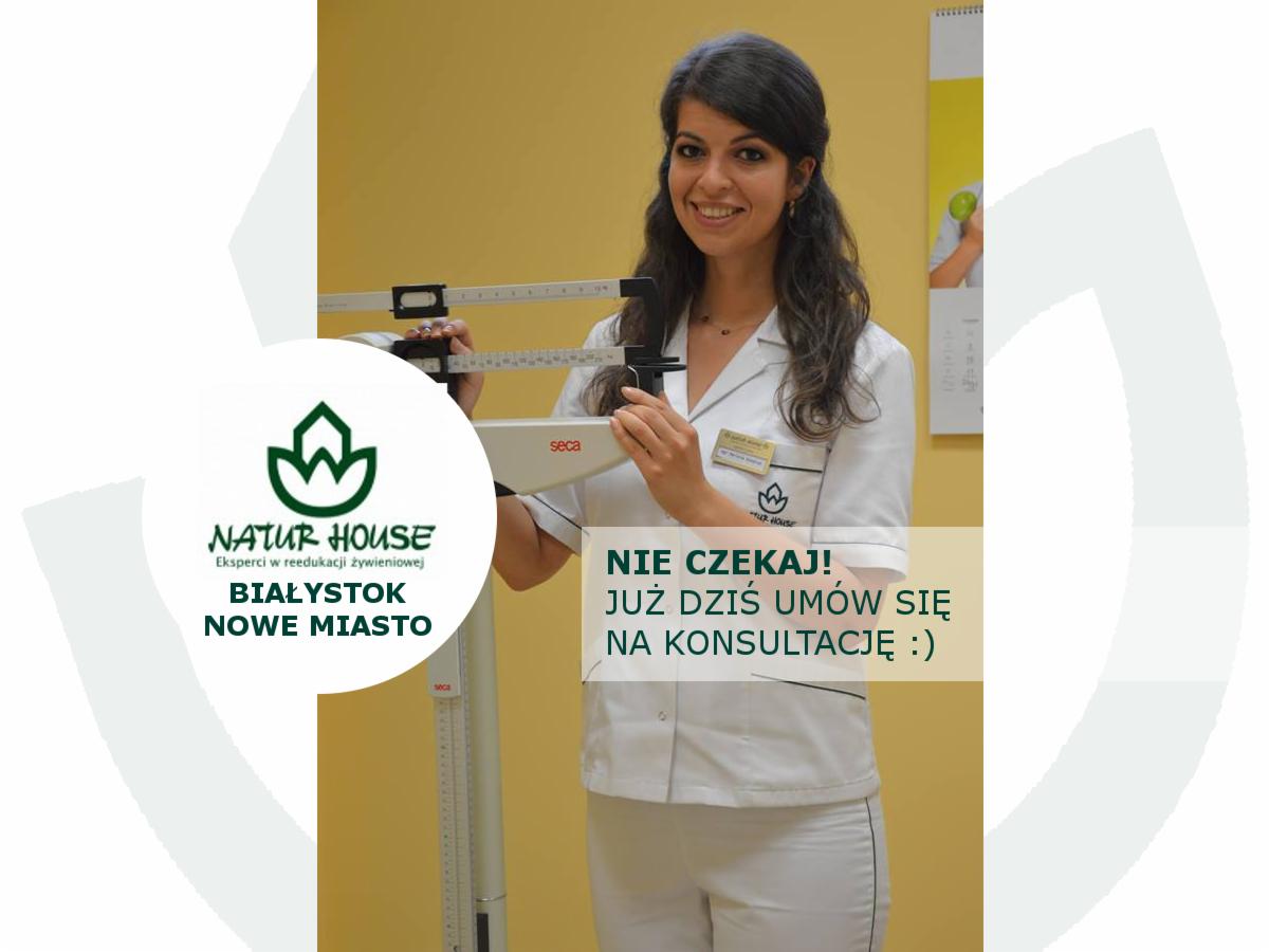 Uśmiechnięta kobieta w białym uniformie medycznym, stojąca obok wagi medycznej marki Seca. Na uniformie widoczna plakietka z logo Natur House. W tle żółta ściana i kalendarz.